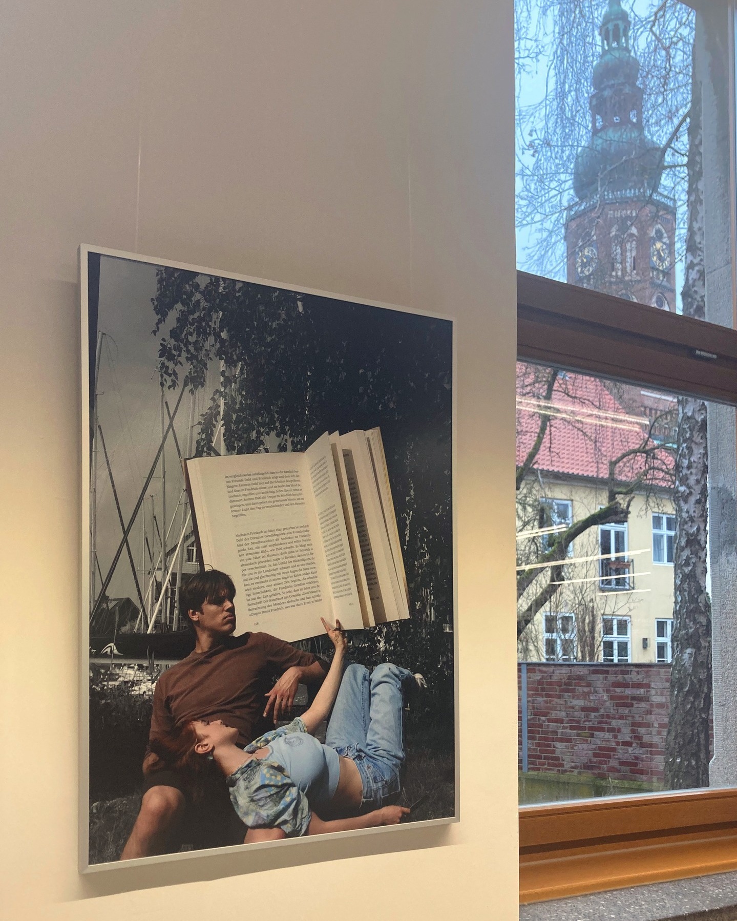 Aufnahme einer ausgestellten Fotografie. Zwei junge Menschen liegen am Hafen, eine Person hält ein übergroßes Buch in der Hand.